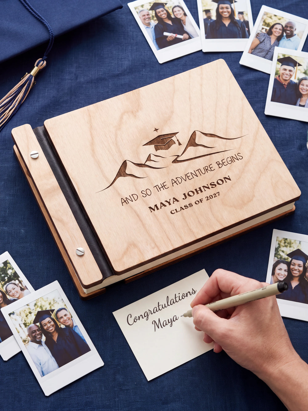 A hand writing a small gift card beside the closed Natural wood graduation photo guest book; the engraved cover and silver screws are visible