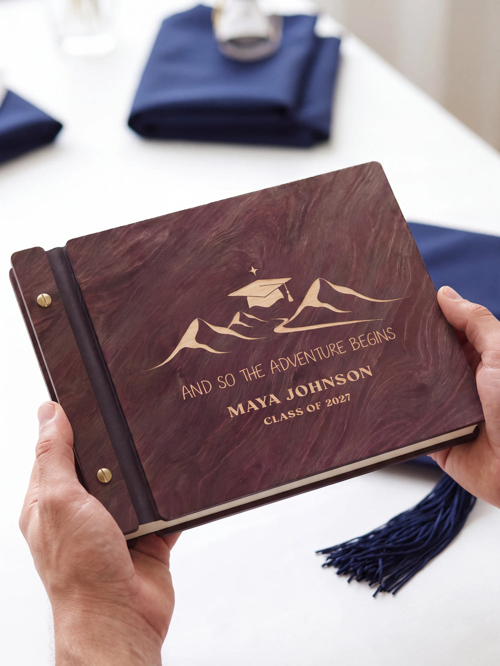 Two hands presenting the closed Jacobean graduation photo guest book with its cream engraving of mountain peaks beneath a graduation cap, a graduation tassel beside them