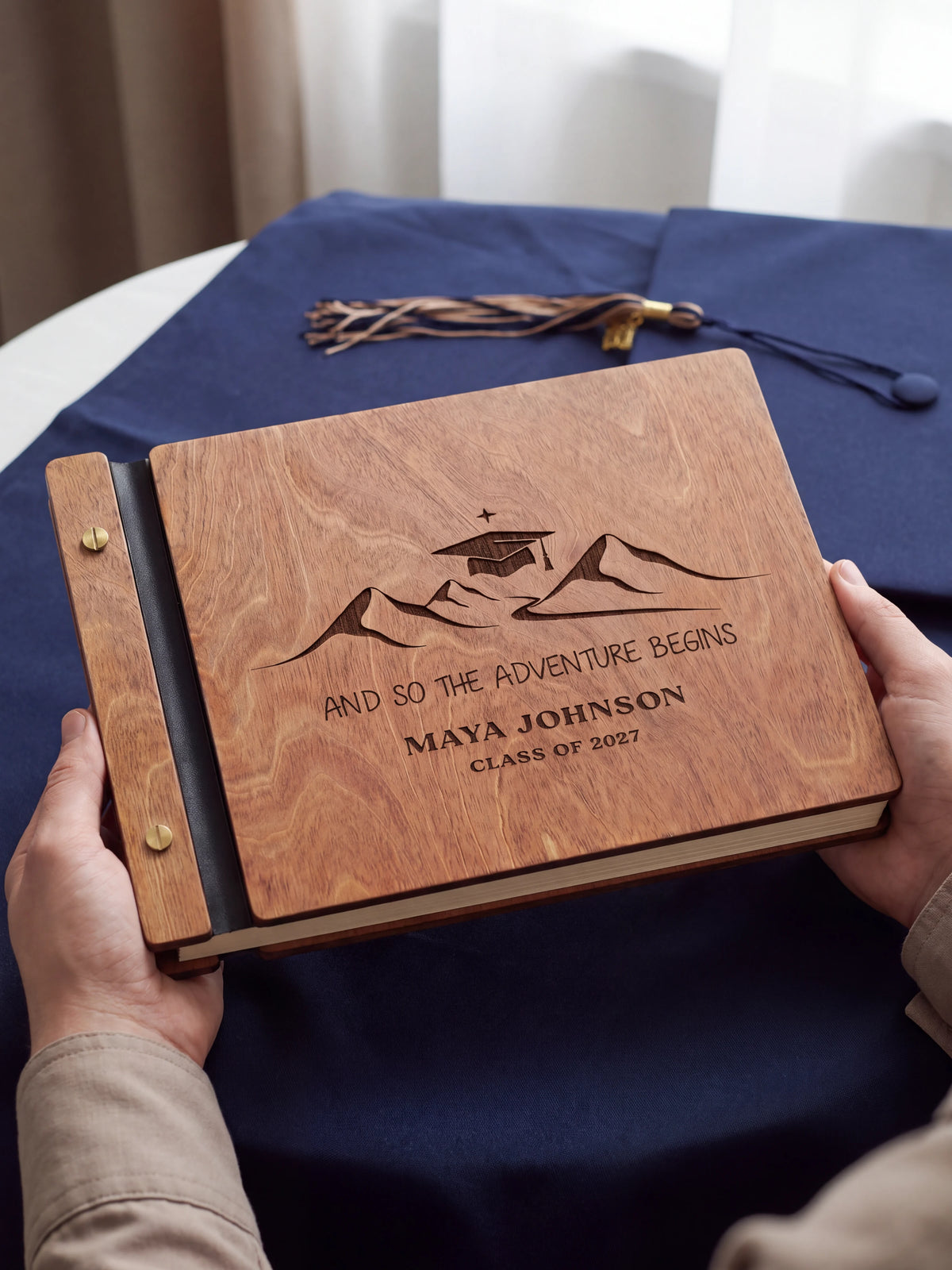 Two hands holding the closed Hazelnut wood graduation photo guest book up to the camera, the dark engraving of mountain peaks beneath a graduation cap and “AND SO THE ADVENTURE BEGINS”, “MAYA JOHNSON” and “CLASS OF 2027” on the lighter wood grain