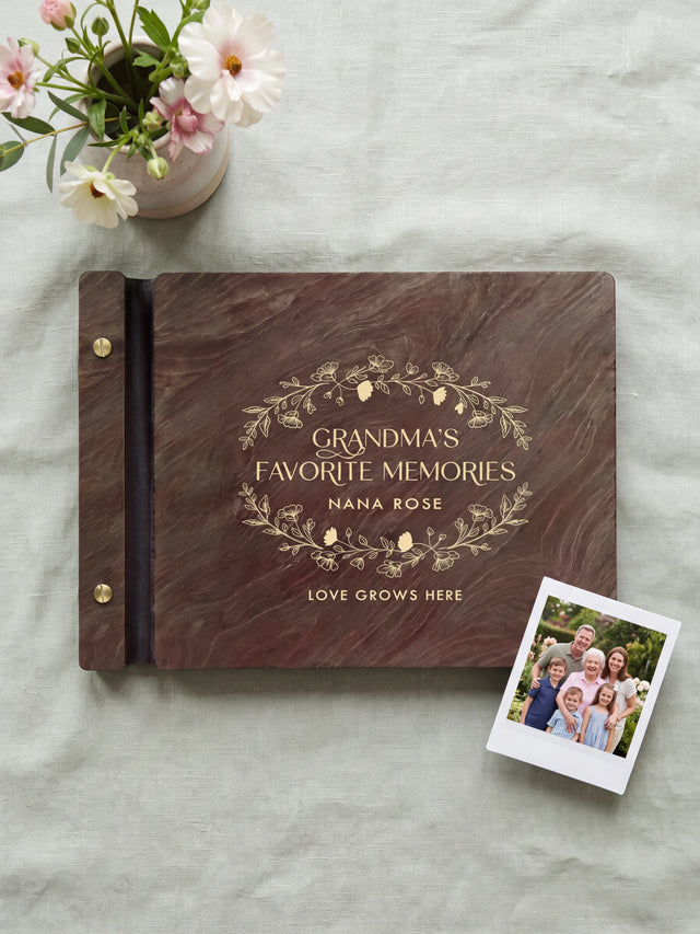 Jacobean wood Grandma's Favorite Memories photo book lying flat on pale sage linen, the cover engraved with “GRANDMA'S FAVORITE MEMORIES”, “NANA ROSE” and “LOVE GROWS HERE”; two gold-tone screws along the black vegan leather spine
