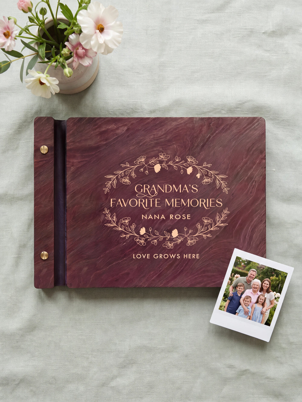 Jacobean wood Grandma's Favorite Memories photo book lying flat on pale sage linen, the cover engraved with “GRANDMA'S FAVORITE MEMORIES”, “NANA ROSE” and “LOVE GROWS HERE”; two gold-tone screws along the black vegan leather spine