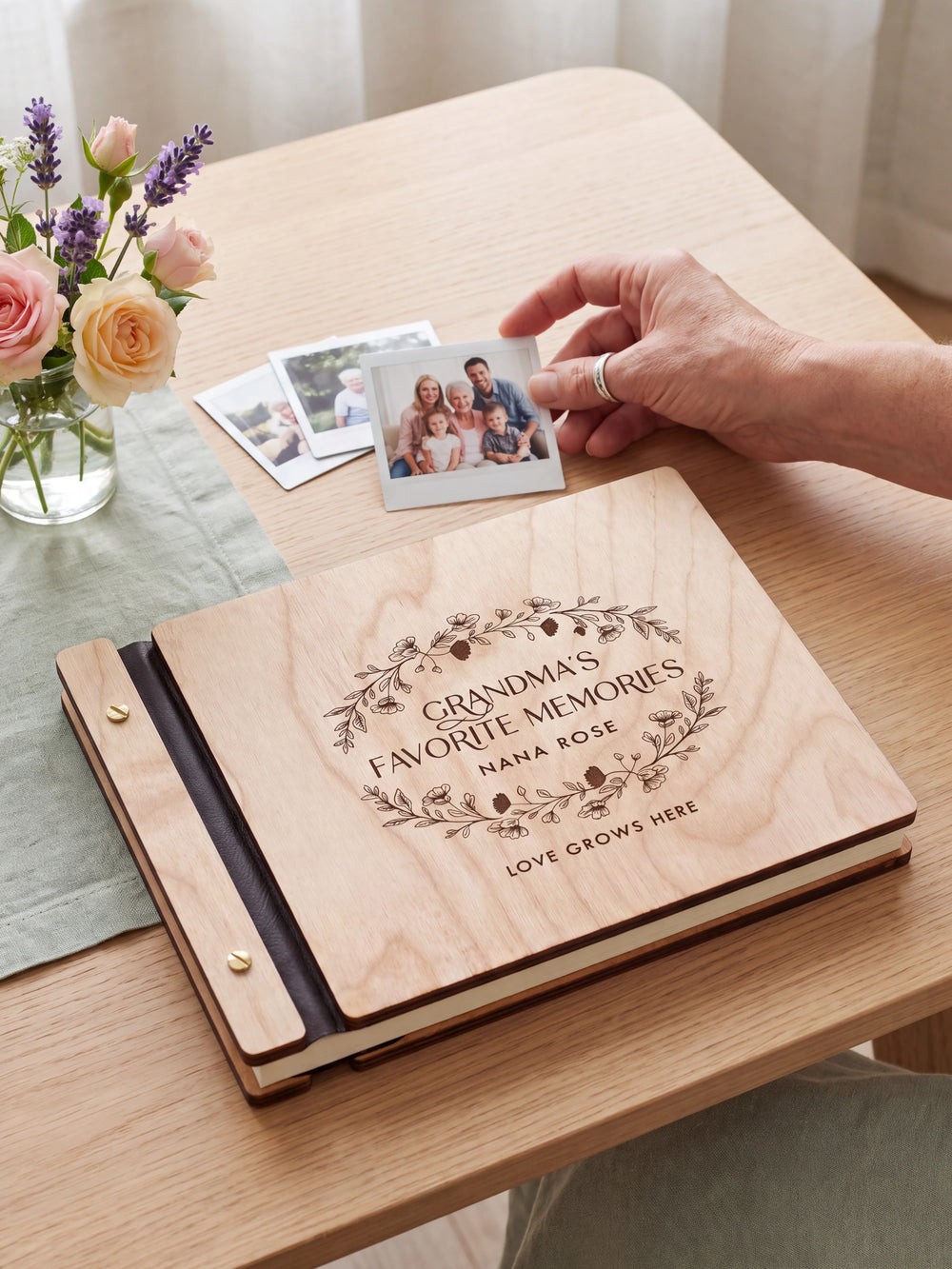 Natural wood Grandma's Favorite Memories photo book on a light table while a hand picks an instant photo to add; the engraved cover reads “GRANDMA'S FAVORITE MEMORIES”, “NANA ROSE” and “LOVE GROWS HERE”