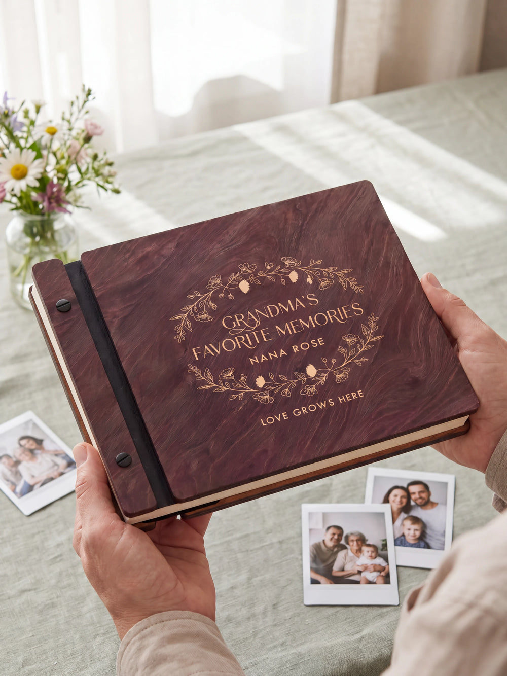 Two hands holding the Jacobean Grandma's Favorite Memories photo book up to the camera, the cream engraving of an oval botanical wreath with small berries and “GRANDMA'S FAVORITE MEMORIES”, “NANA ROSE” and “LOVE GROWS HERE” filling the cover