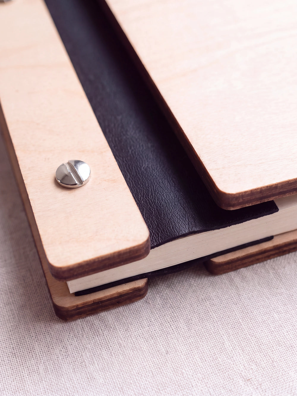 Close-up of a polished silver screw in the Natural wood strip beside the black vegan leather binding, the cream sheet stack visible below