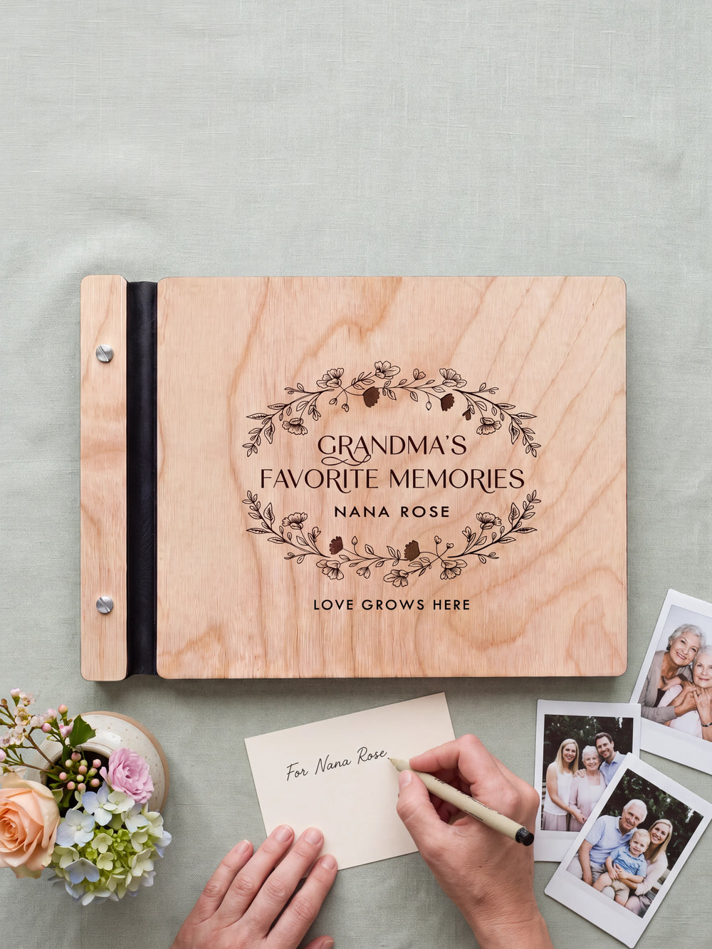 A hand writing a small gift card beside the closed Natural wood Grandma's Favorite Memories photo book; the engraved cover and silver screws are visible