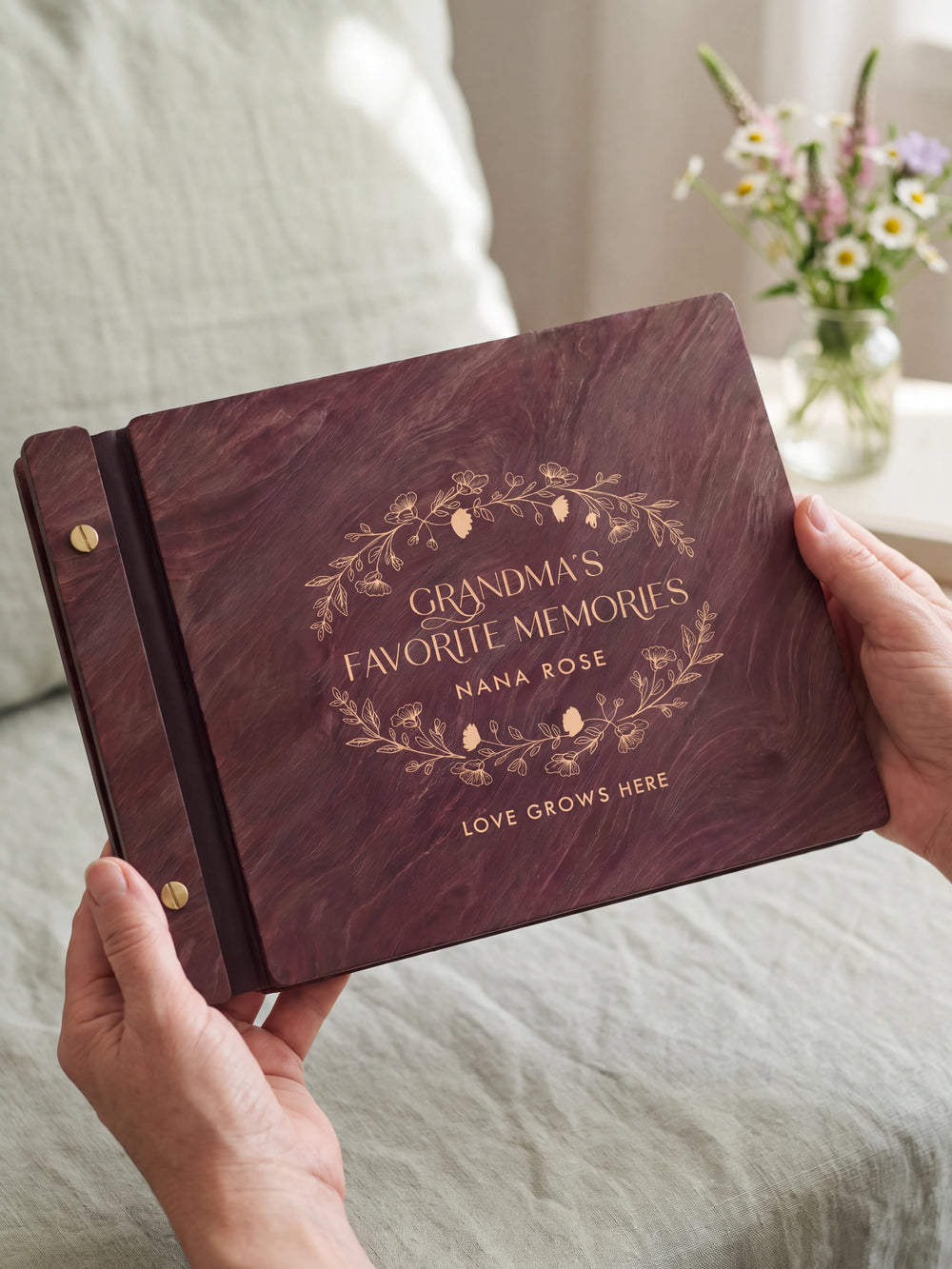 Two hands presenting the closed Jacobean Grandma's Favorite Memories photo book with its cream engraving of an oval botanical wreath with small berries, a small vase of garden flowers beside them