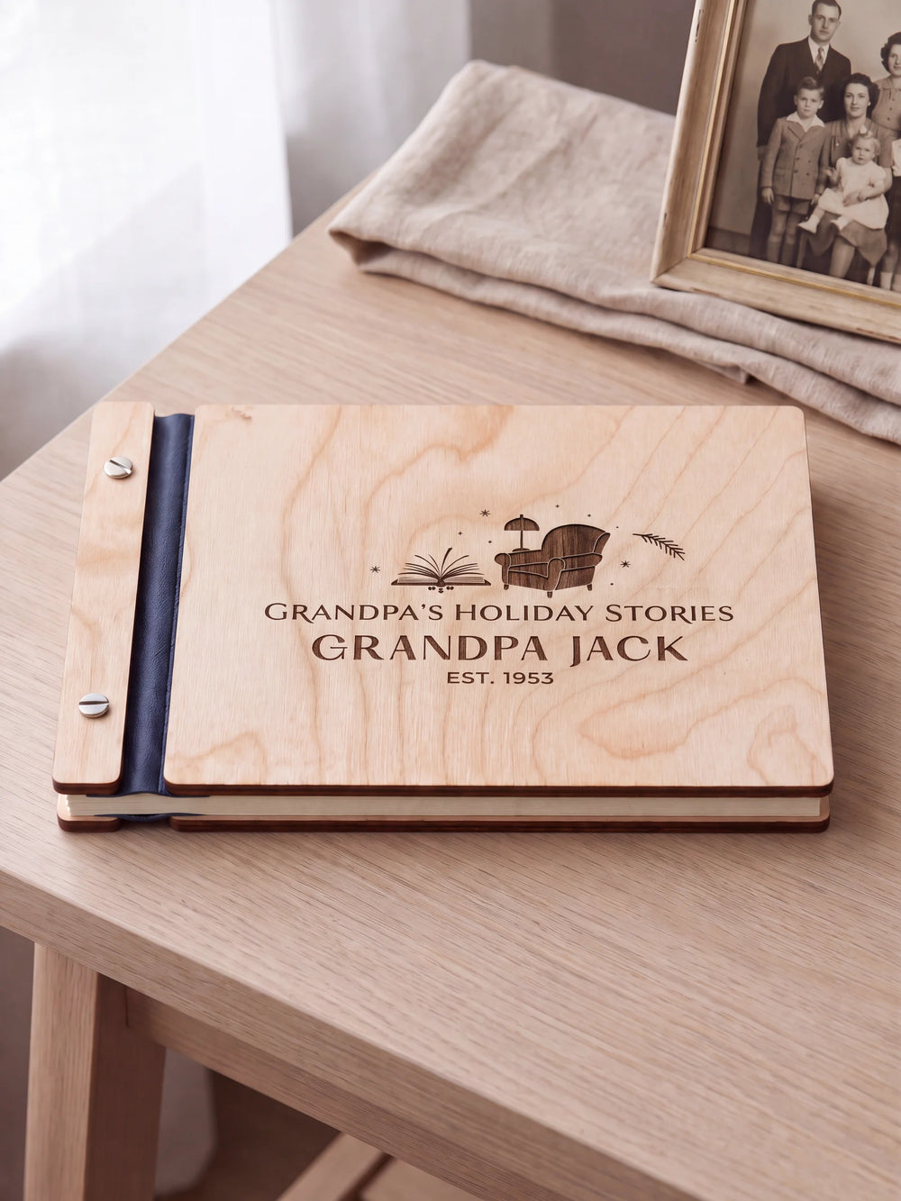 Natural wood Grandpa’s Holiday Stories album on a light wood table beside folded linen and a framed family photograph. The cover has dark brown engraving, navy blue binding and silver screws.