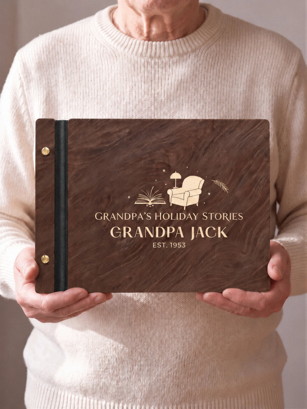 An older man in a cream sweater holds a Jacobean wooden album facing the camera. Light engraved lettering and a reading-chair illustration are visible, with forest green binding and gold screws.