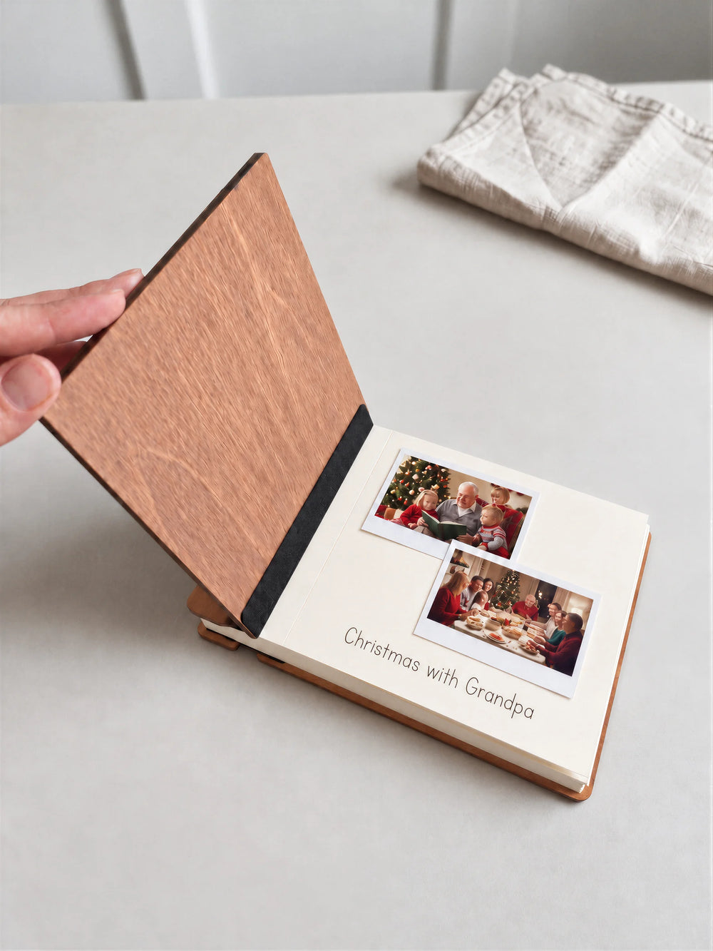 A hand lifts the hazelnut album’s front cover, revealing a black inner fabric strip and a cream page with two family holiday snapshots and the handwritten words Christmas with Grandpa.