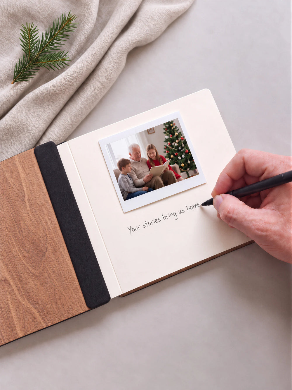 An open hazelnut memory book beside a pine sprig. A hand writes Your stories bring us home below a photograph of a grandfather reading with children; the black inner fabric rests on the wooden cover.