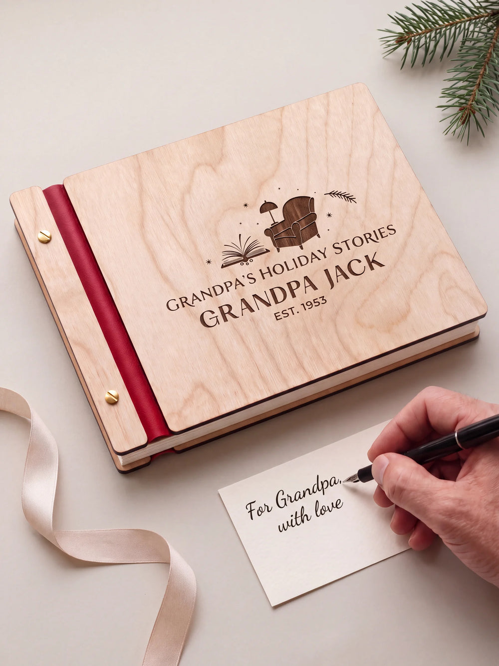 A Natural wood Grandpa’s Holiday Stories album with cherry red binding and gold screws lies beside an ivory ribbon. A hand writes For Grandpa, with love on a small gift card.
