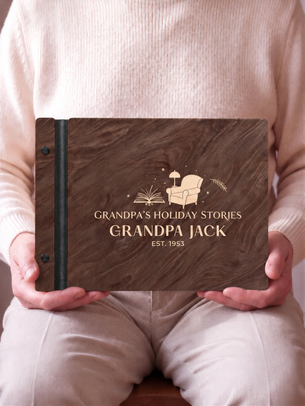 An older man seated in light trousers holds a dark brown Jacobean album on his lap. The cream engraved Grandpa’s Holiday Stories design faces forward, with black binding and matte black screws.