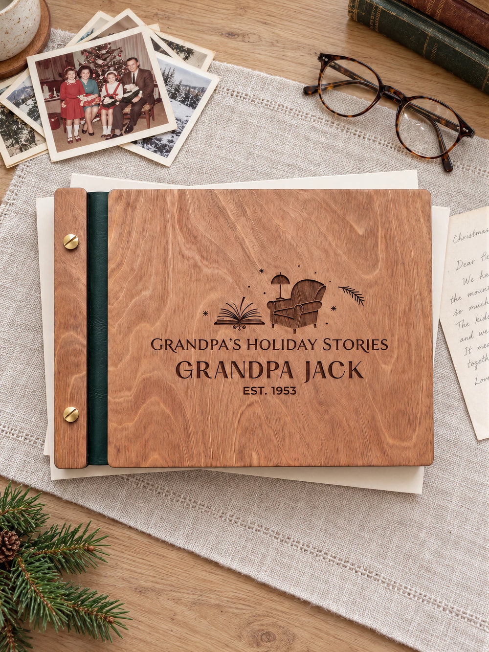 Hazelnut wooden memory book with brown laser engraving, forest green binding and gold screws on a reading table. Old family Christmas photos, glasses and a handwritten letter sit beside the album.
