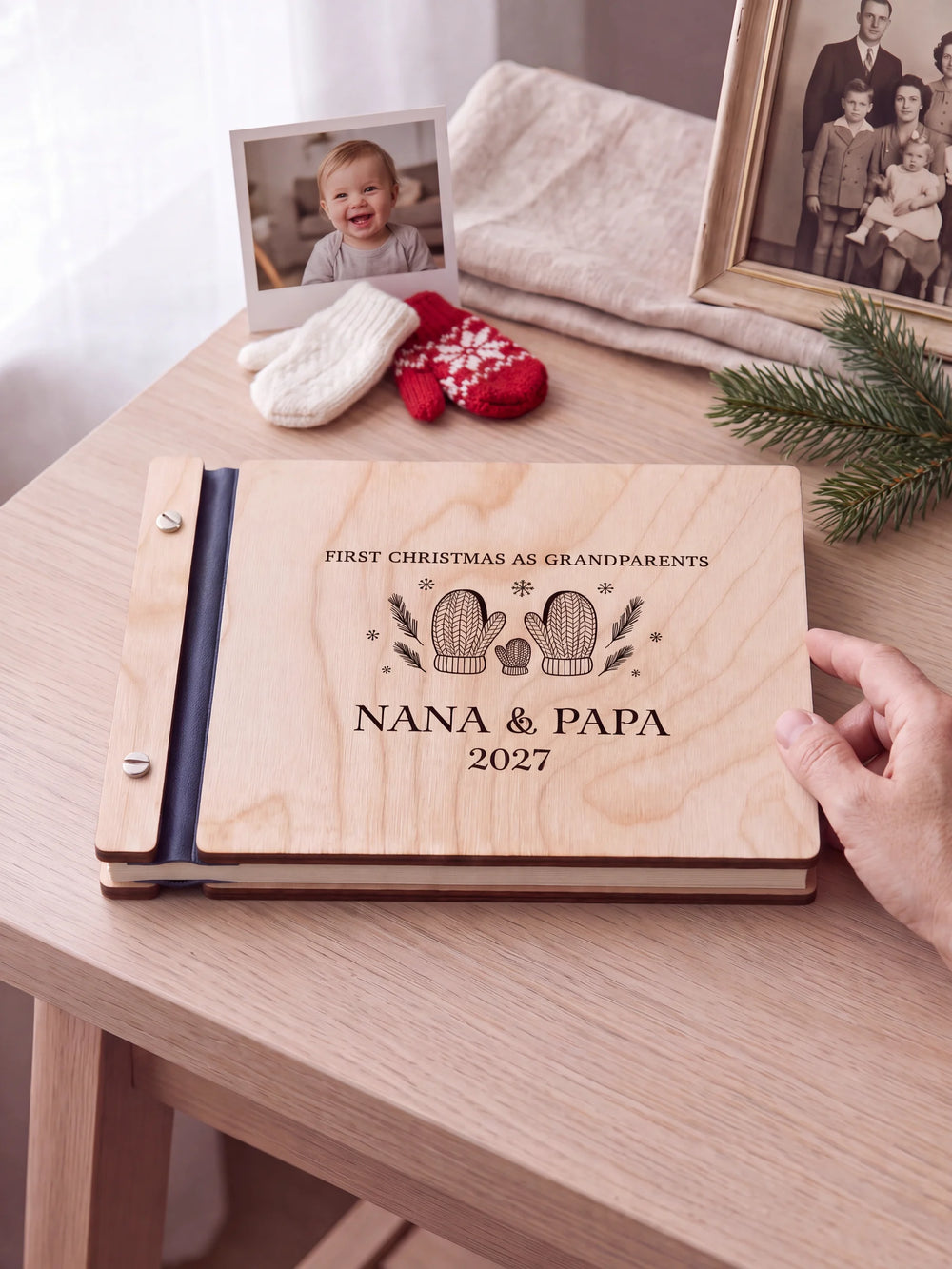 Natural wooden album with brown laser engraving of two knitted mittens and a tiny mitten, shown in a close lifestyle view with navy blue binding and silver screws. The cover reads FIRST CHRISTMAS AS GRANDPARENTS.