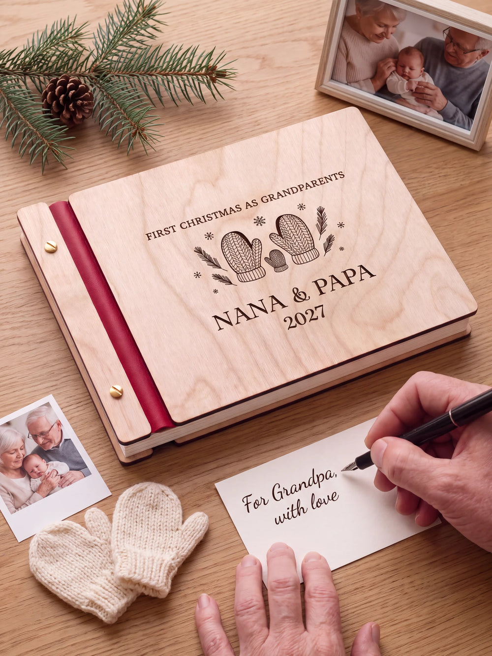 A Natural wooden album with the FIRST CHRISTMAS AS GRANDPARENTS engraving, cherry red binding and gold screws. A hand writes on a separate gift card beside the book.
