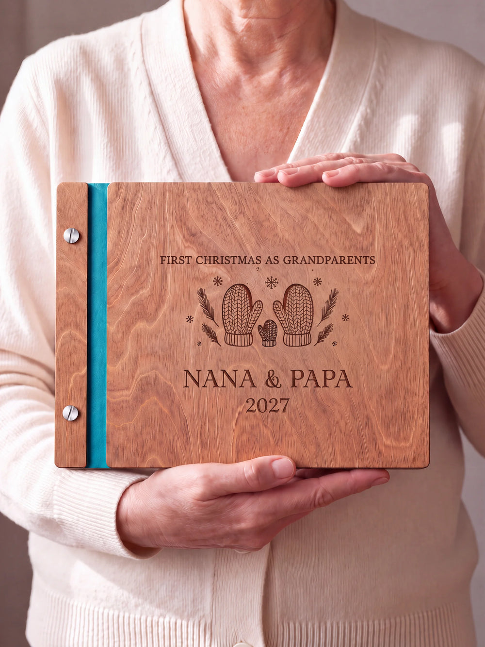 Two hands hold a Hazelnut wooden photo album. The FIRST CHRISTMAS AS GRANDPARENTS cover design has brown engraving, turquoise binding and silver screws.