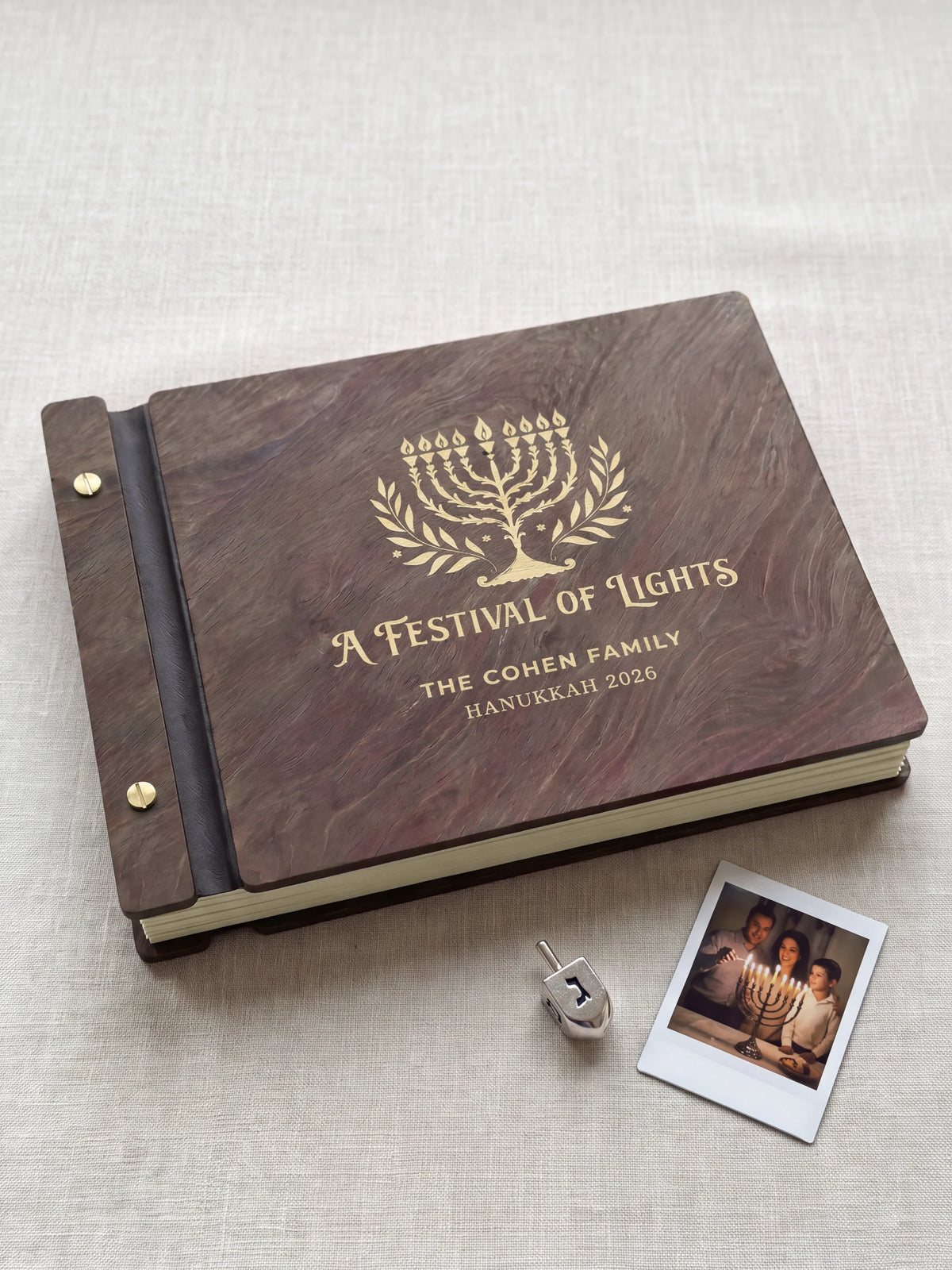Jacobean wood Hanukkah photo memory book lying flat on navy linen, the cover engraved with “A FESTIVAL OF LIGHTS”, “THE COHEN FAMILY” and “HANUKKAH 2026”; two gold-tone screws along the black vegan leather spine