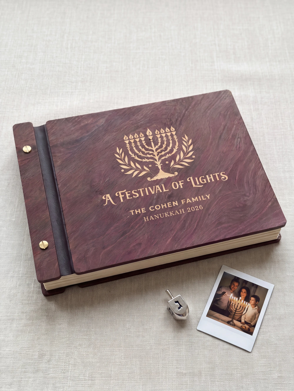 Jacobean wood Hanukkah photo memory book lying flat on navy linen, the cover engraved with “A FESTIVAL OF LIGHTS”, “THE COHEN FAMILY” and “HANUKKAH 2026”; two gold-tone screws along the black vegan leather spine