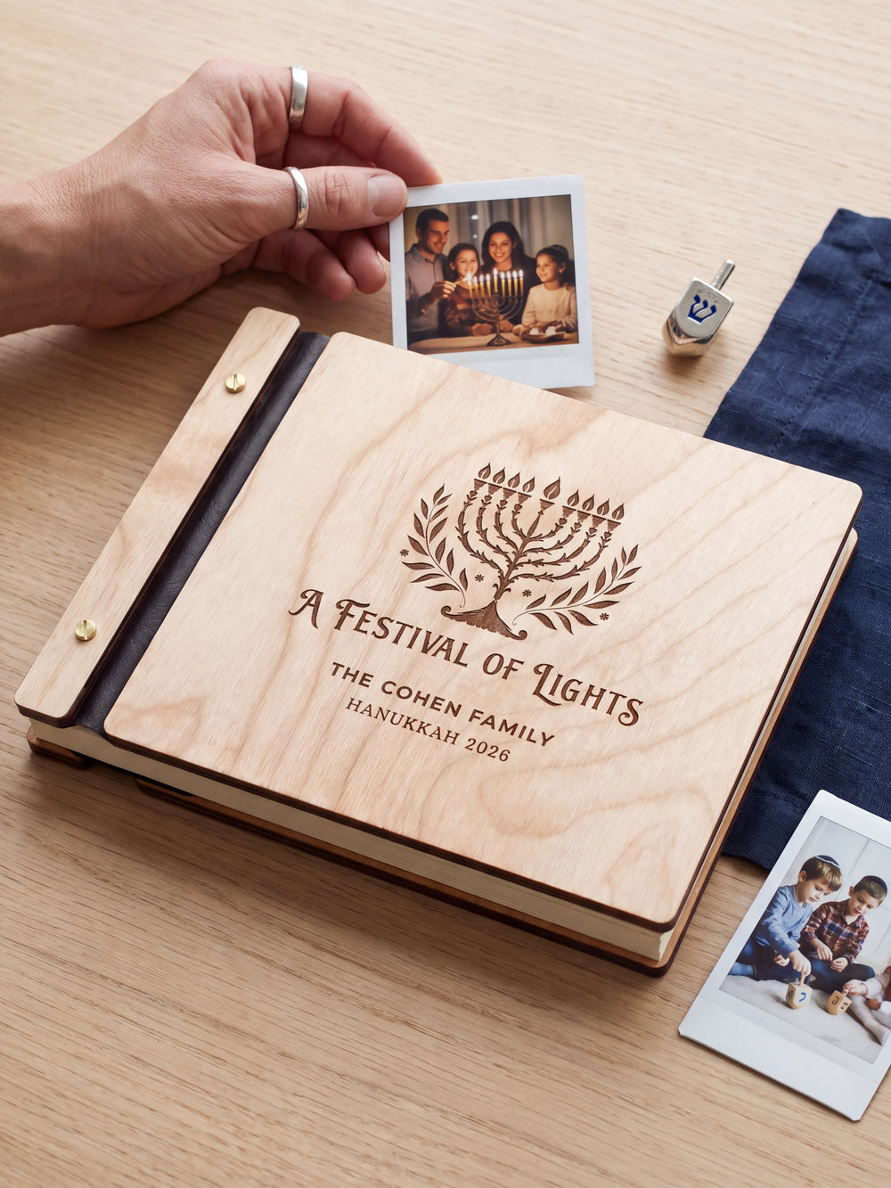 Natural wood Hanukkah photo memory book on a light table while a hand picks an instant photo to add; the engraved cover reads “A FESTIVAL OF LIGHTS”, “THE COHEN FAMILY” and “HANUKKAH 2026”