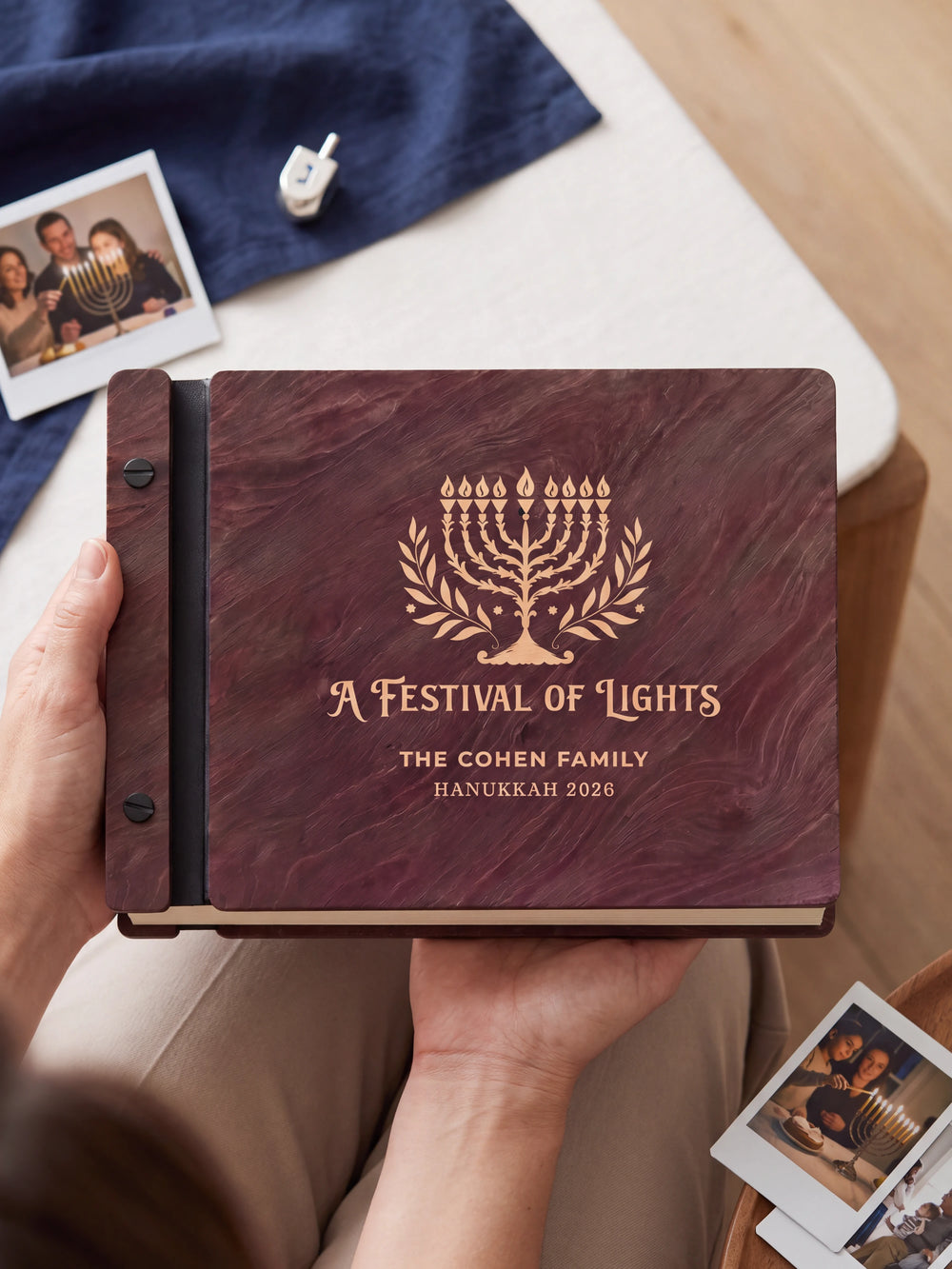 Two hands holding the Jacobean Hanukkah photo memory book up to the camera, the cream engraving of a nine-branched menorah with leafy branches and “A FESTIVAL OF LIGHTS”, “THE COHEN FAMILY” and “HANUKKAH 2026” filling the cover
