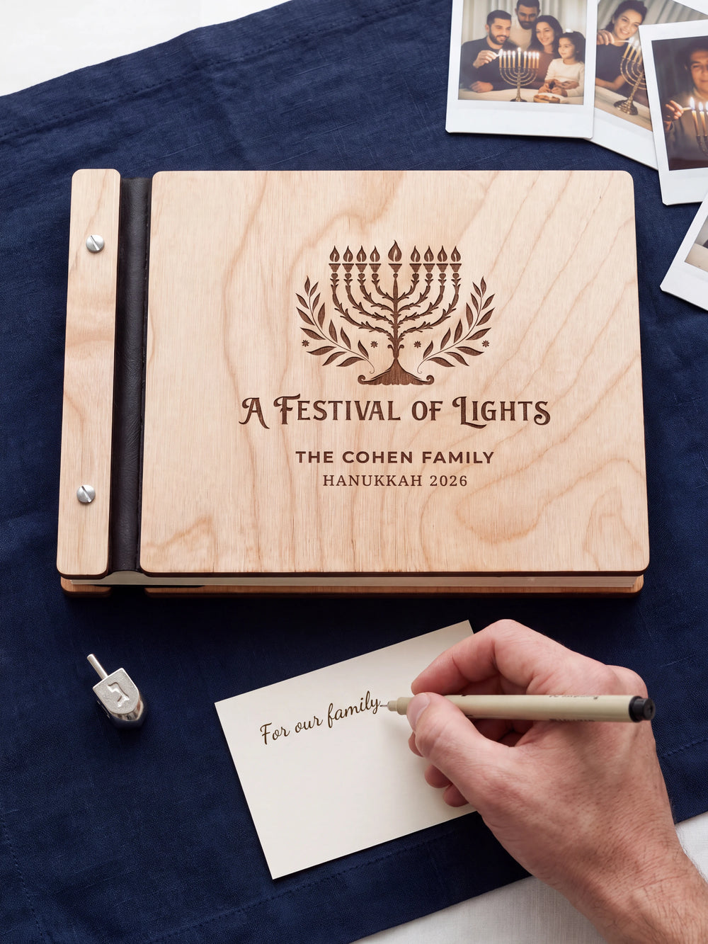 A hand writing a small gift card beside the closed Natural wood Hanukkah photo memory book; the engraved cover and silver screws are visible