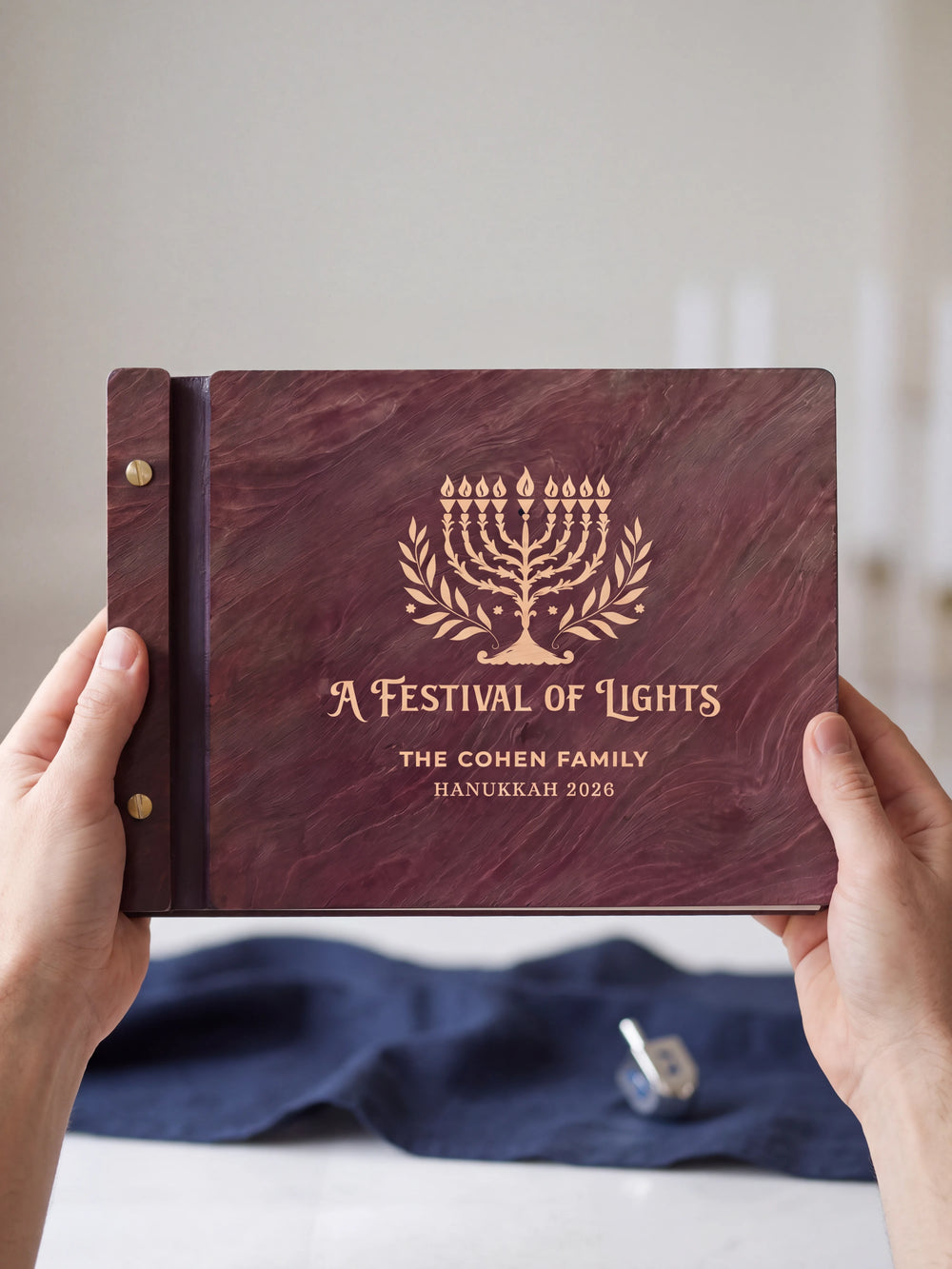 Two hands presenting the closed Jacobean Hanukkah photo memory book with its cream engraving of a nine-branched menorah with leafy branches, a small silver dreidel on the table beside them