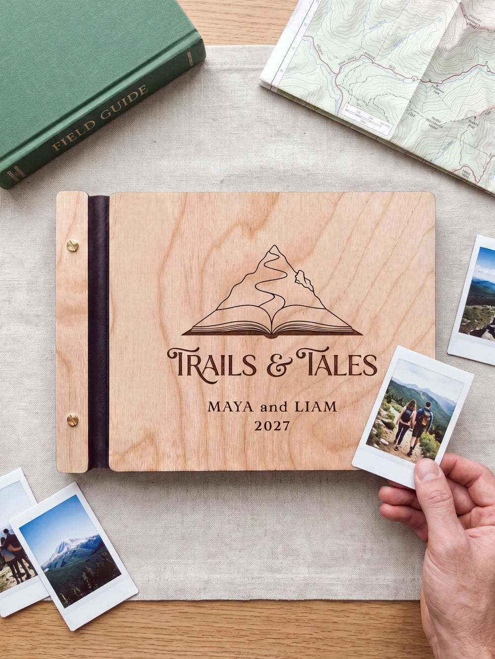 Natural wooden album with dark engraving of a mountain rising from an open book. A hand holds an instant photo beside the closed cover, with loose snapshots nearby.
