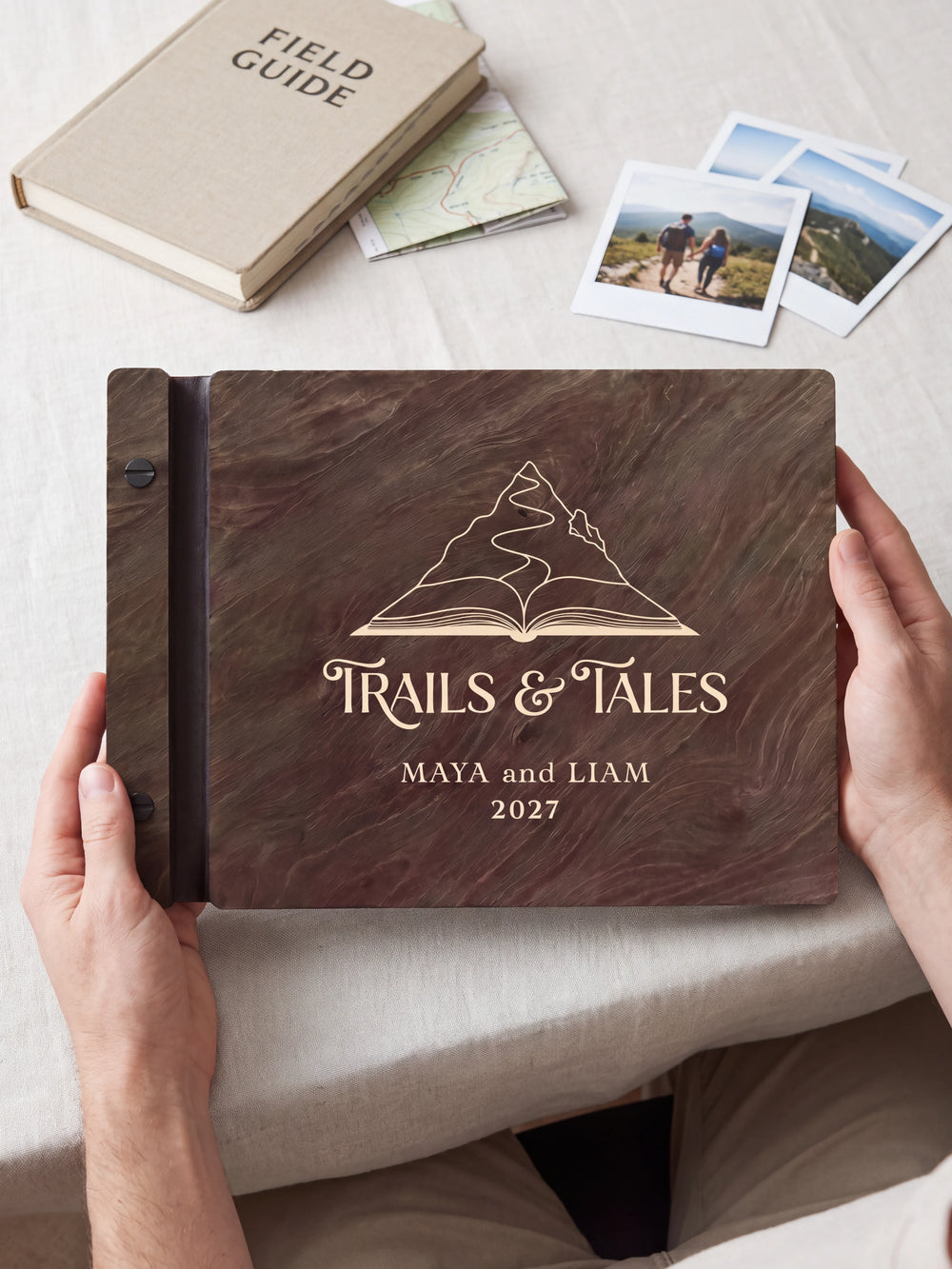 Hands hold a dark brown Jacobean wooden album close to the camera. Cream engraving shows a mountain rising from an open book, personalized wording and the black binding strip.