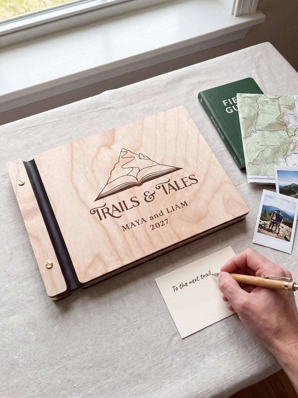 A Natural wooden album showing a mountain rising from an open book rests beside a separate gift card. A hand writes 
