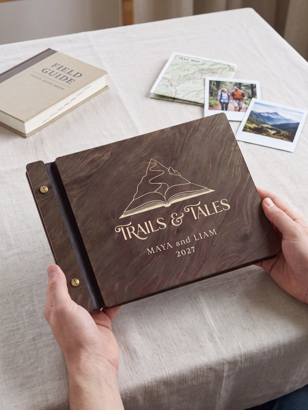 Two hands present a dark brown Jacobean wooden album, with the full cream-engraved mountain rising from an open book design and personalized cover wording facing the camera.