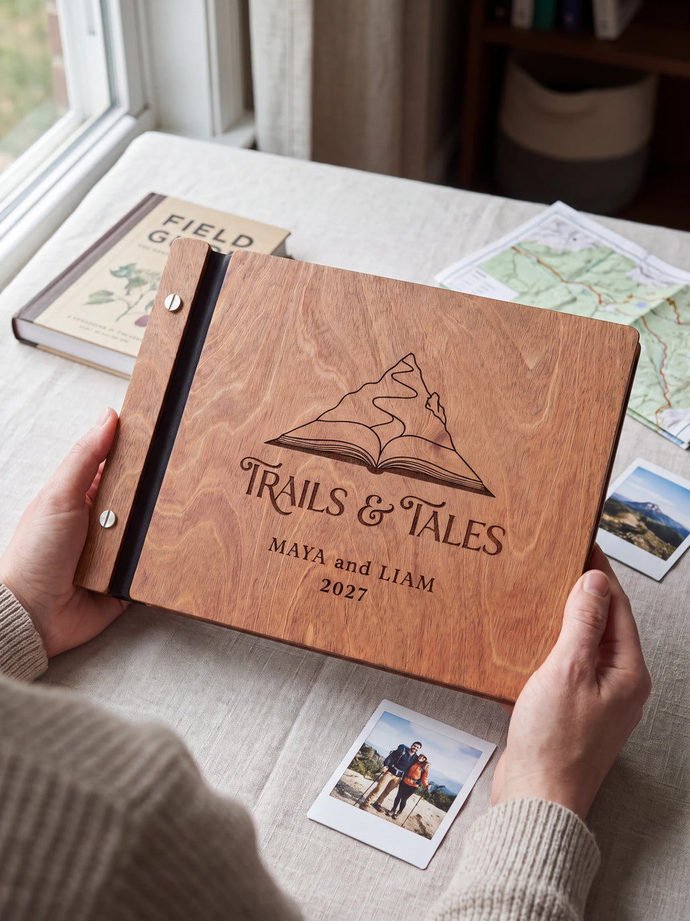 Hands hold the Hazelnut wooden album near a window. The full cover displays a mountain rising from an open book in dark engraving, with personalized lettering and a black binding strip.