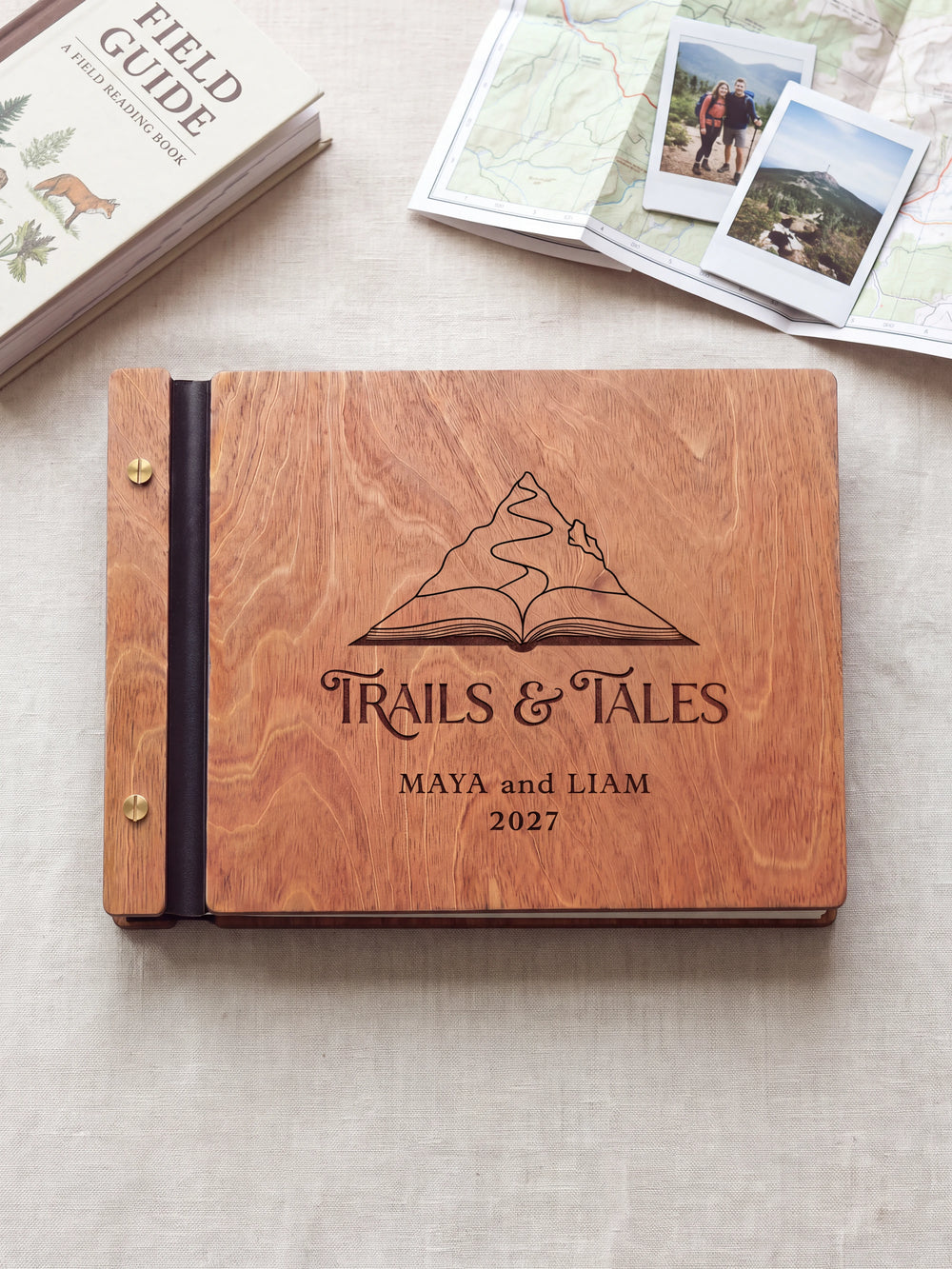 Hazelnut wooden album on a light tabletop with neutral linen. The full cover shows a mountain rising from an open book, personalized wording and the two-screw binding.
