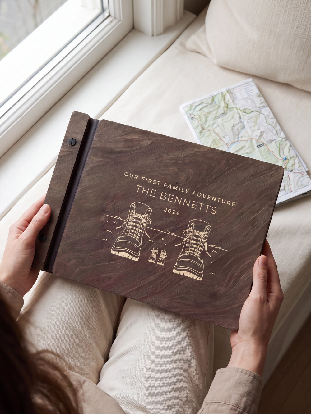 Hands hold a dark brown Jacobean wooden album close to the camera. Cream engraving shows adult and little hiking boots, personalized wording and the black binding strip.