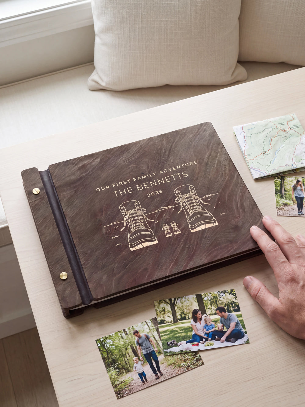 A hand rests beside the closed dark brown Jacobean album on a table. Cream engraving and personalized wording are visible, with travel photos nearby.