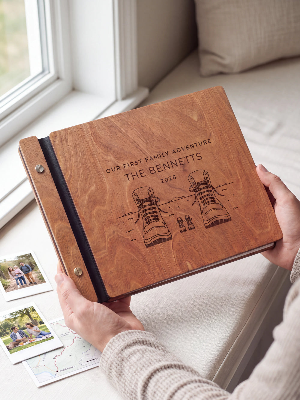 Hands hold the Hazelnut wooden album near a window. The full cover displays adult and little hiking boots in dark engraving, with personalized lettering and a black binding strip.