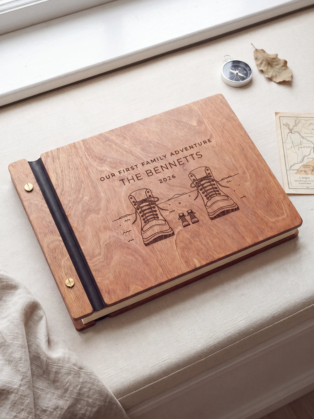 Hazelnut wooden album on a light tabletop with neutral linen. The full cover shows adult and little hiking boots, personalized wording and the two-screw binding.