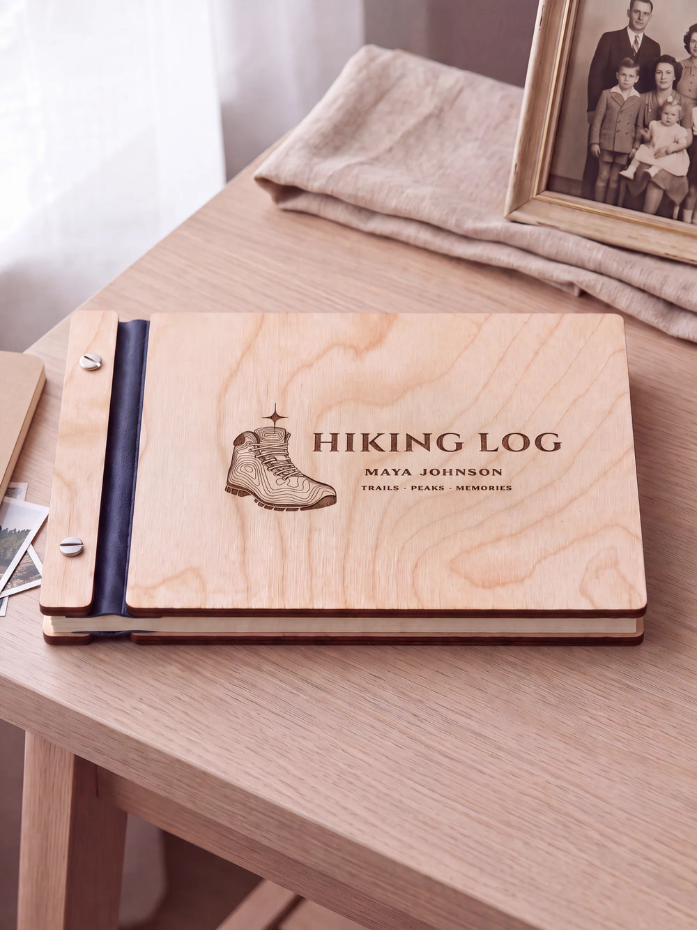 Natural wooden album with brown laser engraving of hiking boot with topographic contours and star, shown in a close lifestyle view with navy blue binding and silver screws. The cover reads HIKING LOG.