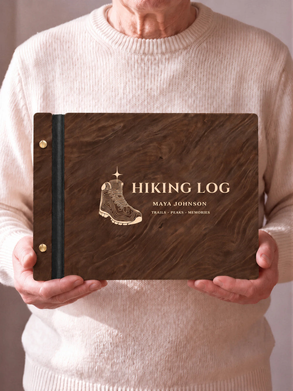 Two hands hold a Jacobean wooden photo album with the HIKING LOG cover facing the camera. Cream engraved artwork, the narrow binding and gold screws are visible.