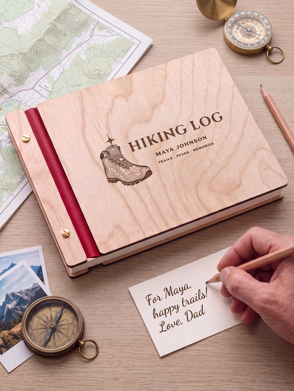 A Natural wooden album with the HIKING LOG engraving, cherry red binding and gold screws. A hand writes on a separate gift card beside the book.