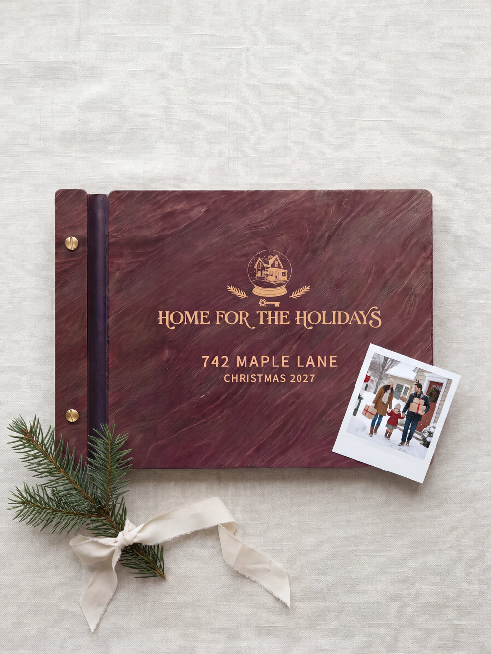 Jacobean wood Home for the Holidays photo book lying flat on a light entry table, the cover engraved with “HOME FOR THE HOLIDAYS”, “742 MAPLE LANE” and “CHRISTMAS 2027”; two gold-tone screws along the black vegan leather spine