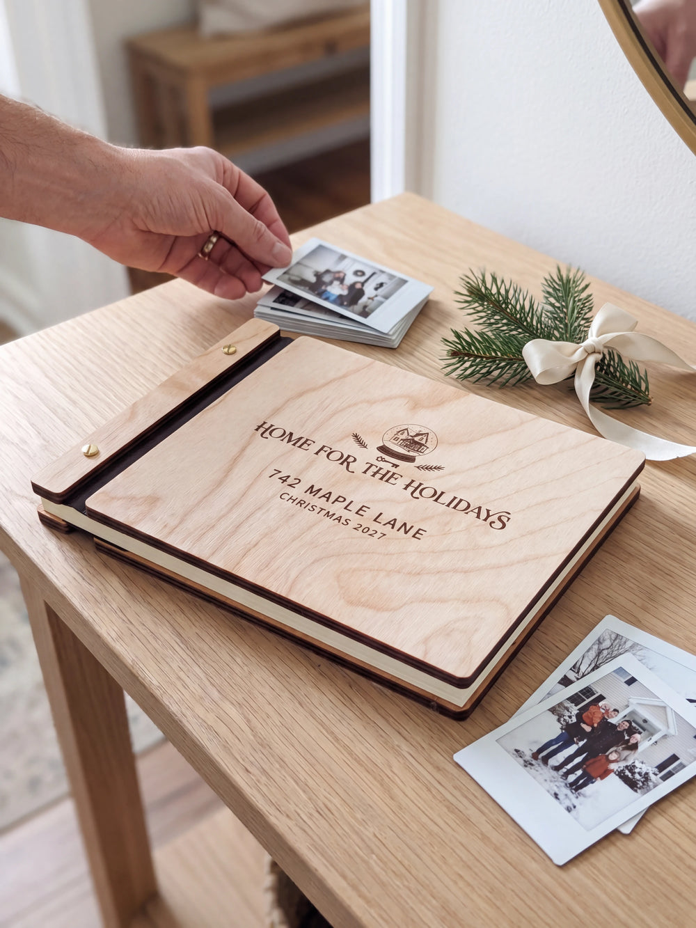 Natural wood Home for the Holidays photo book on a light table while a hand picks an instant photo to add; the engraved cover reads “HOME FOR THE HOLIDAYS”, “742 MAPLE LANE” and “CHRISTMAS 2027”