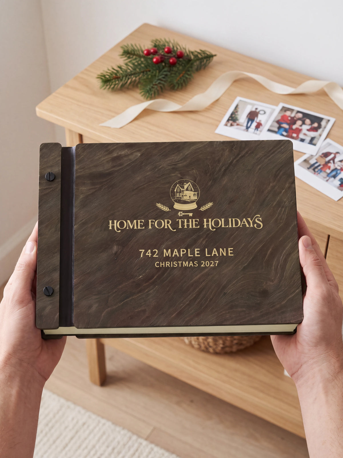 Two hands holding the Jacobean Home for the Holidays photo book up to the camera, the gold-tone engraving of a winter house inside a snow globe with evergreen sprigs and “HOME FOR THE HOLIDAYS”, “742 MAPLE LANE” and “CHRISTMAS 2027” filling the cover