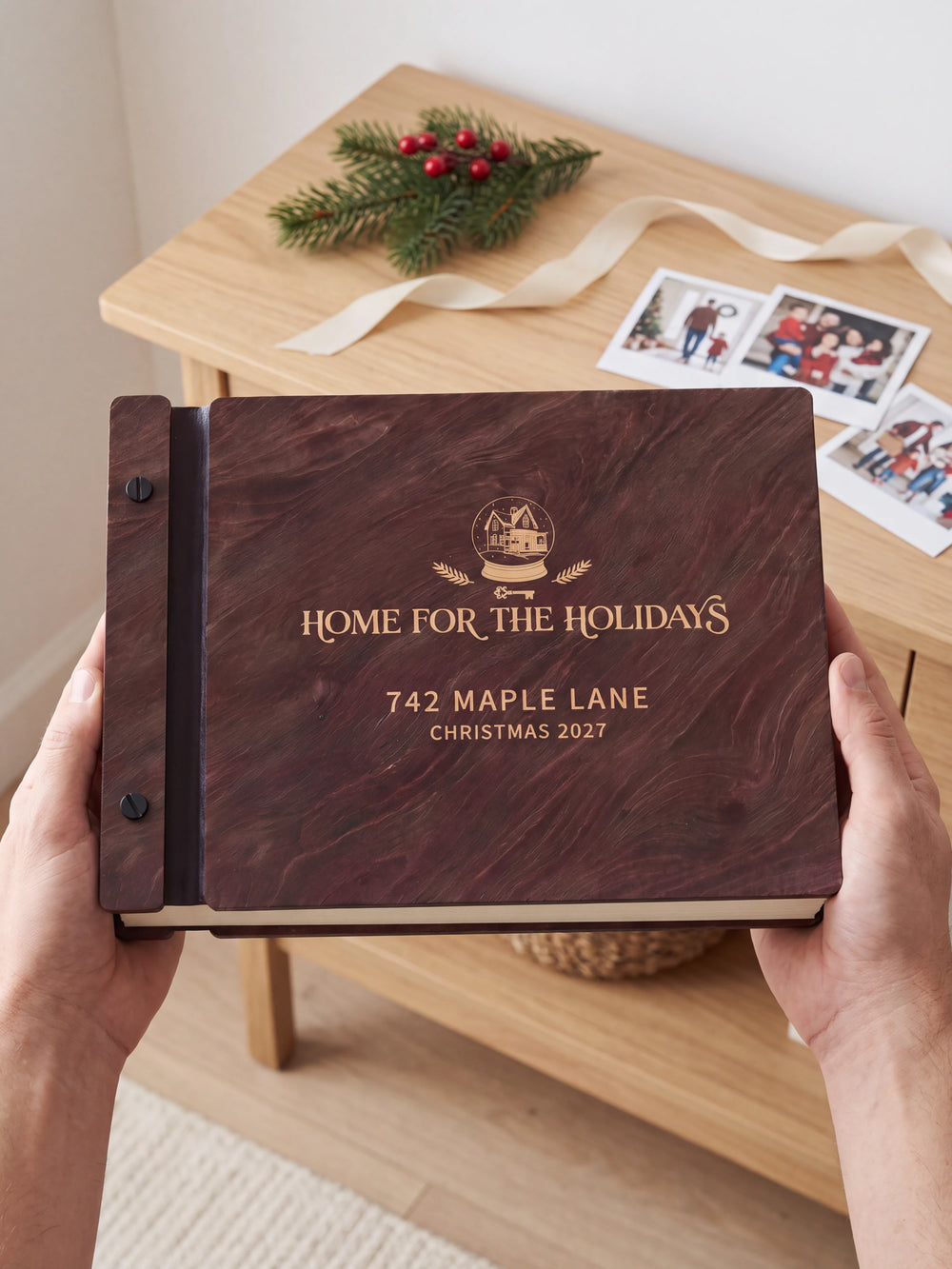 Two hands holding the Jacobean Home for the Holidays photo book up to the camera, the cream engraving of a winter house inside a snow globe with evergreen sprigs and “HOME FOR THE HOLIDAYS”, “742 MAPLE LANE” and “CHRISTMAS 2027” filling the cover