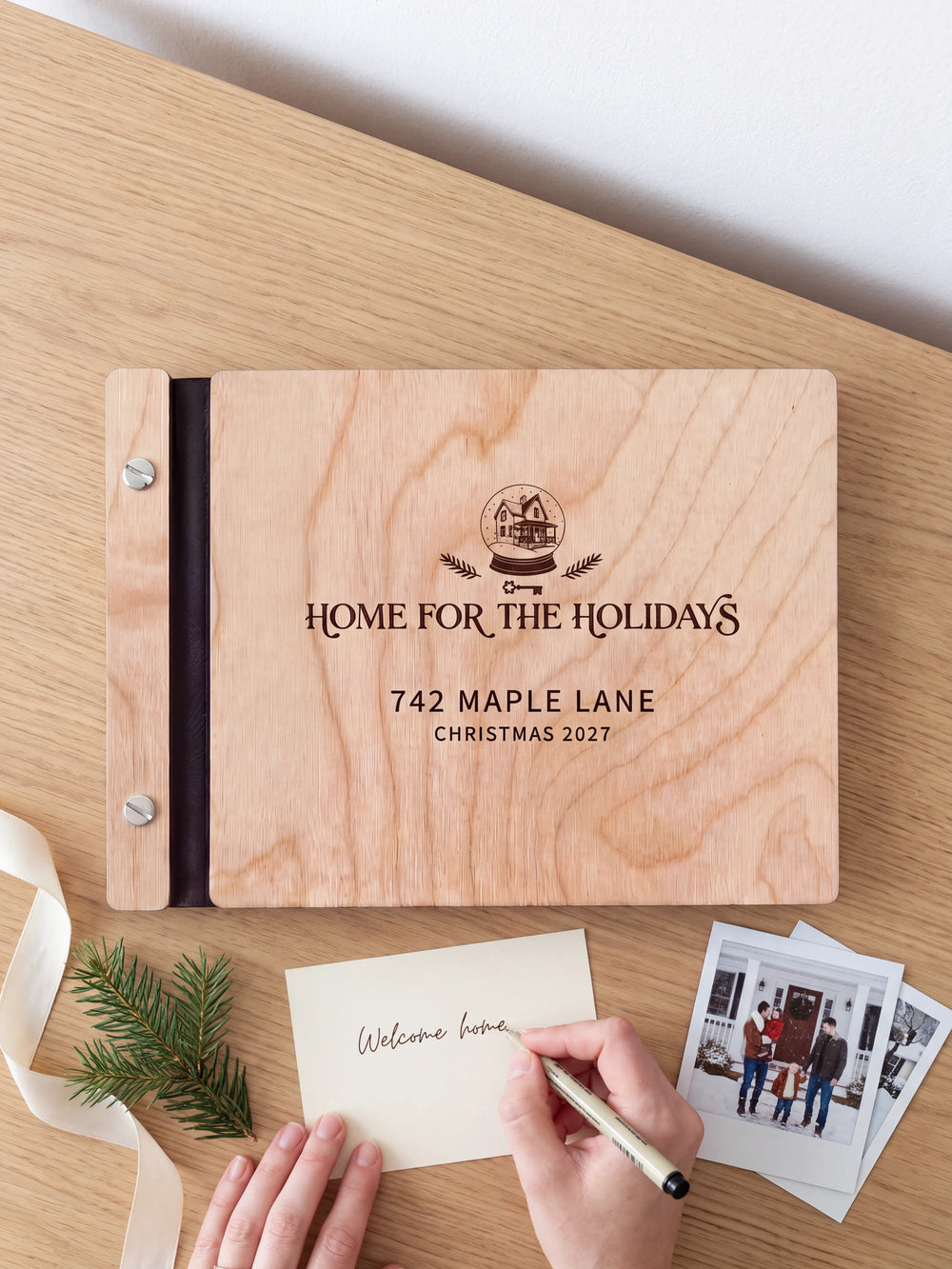A hand writing a small gift card beside the closed Natural wood Home for the Holidays photo book; the engraved cover and silver screws are visible