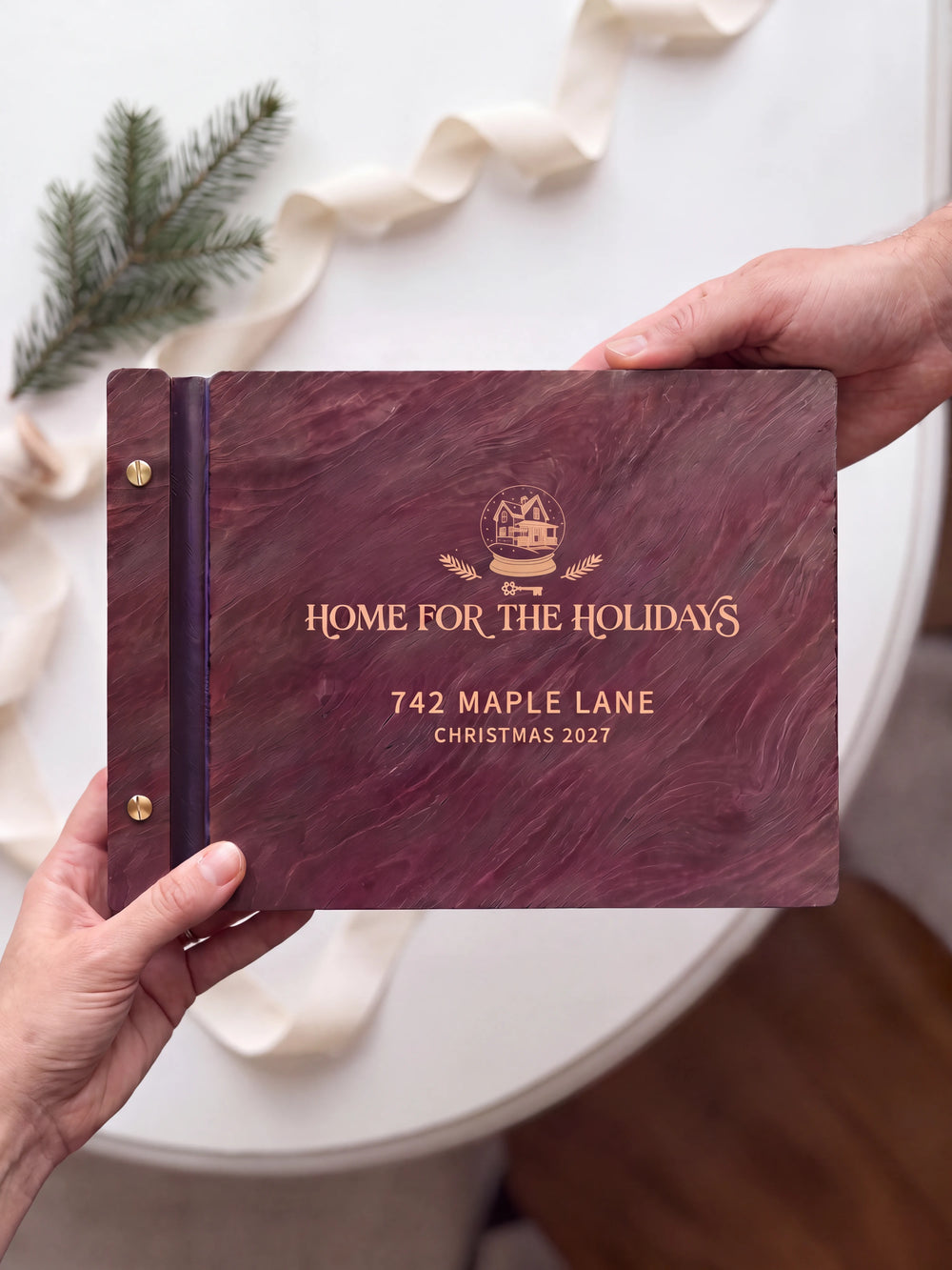 Two hands presenting the closed Jacobean Home for the Holidays photo book with its cream engraving of a winter house inside a snow globe with evergreen sprigs, an evergreen sprig and ivory ribbon on the table beside them
