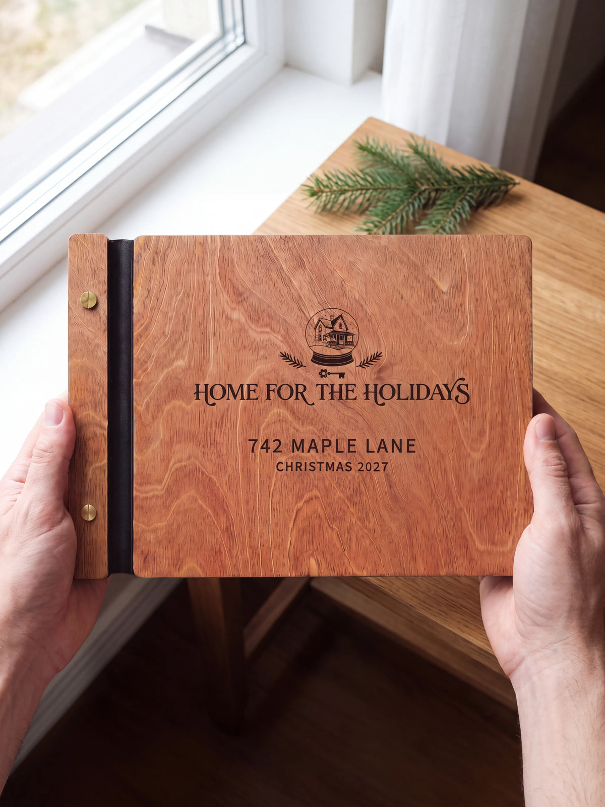 Two hands holding the closed Hazelnut wood Home for the Holidays photo book up to the camera, the dark engraving of a winter house inside a snow globe with evergreen sprigs and “HOME FOR THE HOLIDAYS”, “742 MAPLE LANE” and “CHRISTMAS 2027” on the lighter wood grain