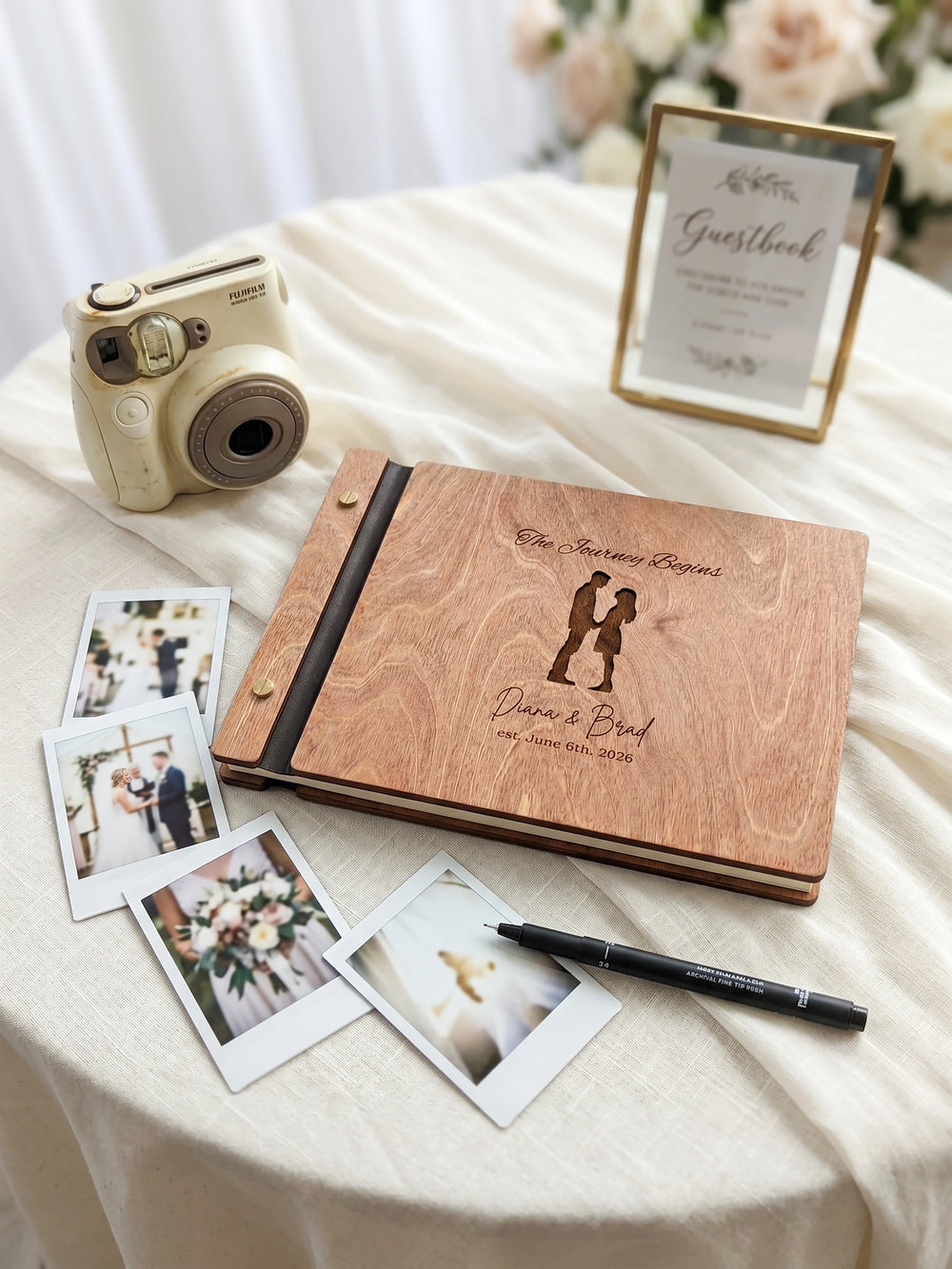 Hazelnut wood The Journey Begins photo guest book engraved with a couple silhouette, Diana & Brad est. June 6th, 2026, on a round table with an instant camera and photos