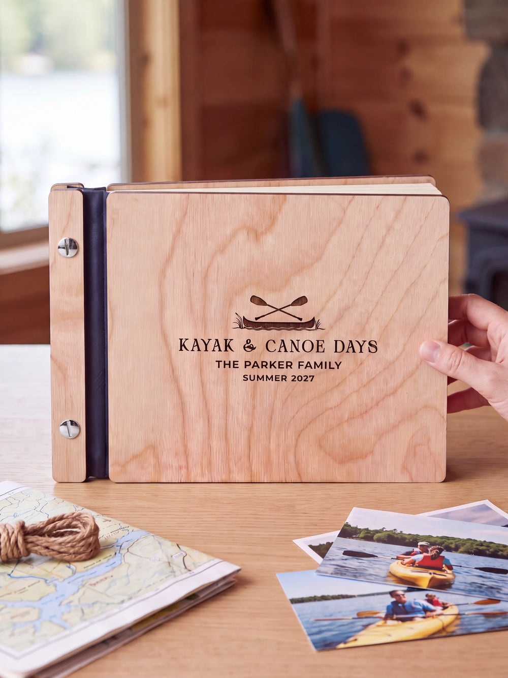 Natural wooden album with brown laser engraving of canoe and crossed paddles, shown in a close lifestyle view with navy blue binding and silver screws. The cover reads KAYAK & CANOE DAYS.