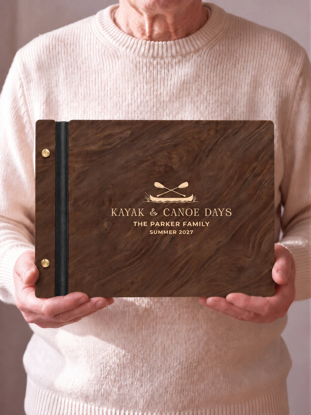 Two hands hold a Jacobean wooden photo album with the KAYAK & CANOE DAYS cover facing the camera. Cream engraved artwork, the narrow binding and gold screws are visible.