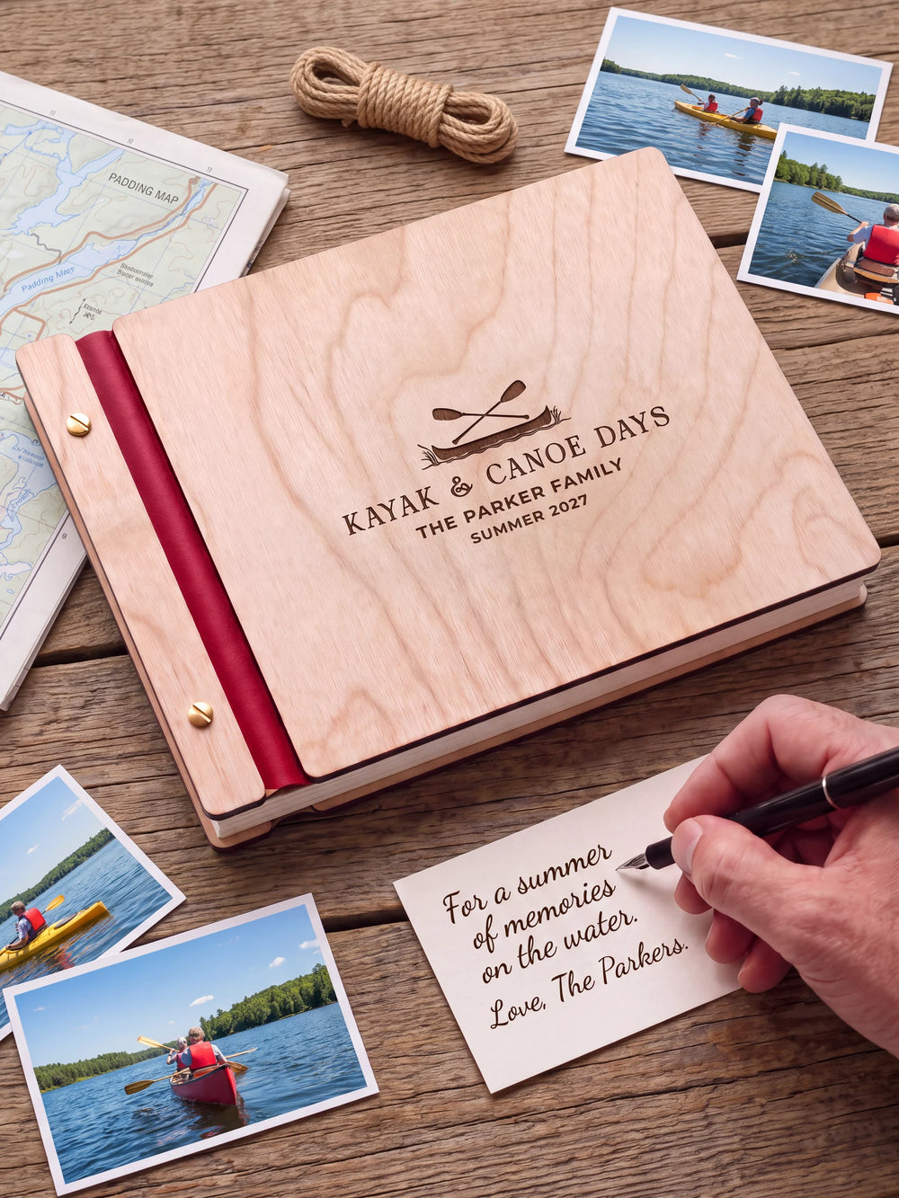 A Natural wooden album with the KAYAK & CANOE DAYS engraving, cherry red binding and gold screws. A hand writes on a separate gift card beside the book.